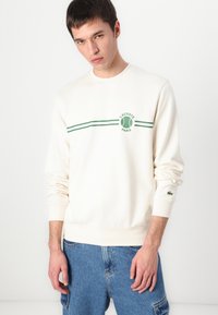 Cream sweatshirt made of cotton, featuring green stripes and a tennis ball logo. Ribbed cuffs and hem with Lacoste crocodile accent on sleeve.
