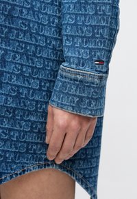 Keskitummansininen farkkumekko, jossa on koko pinnan kattava logokuvio, ommellut yksityiskohdat ja epäsymmetrinen helma.