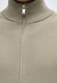 Gros plan d'une veste en maille beige texturée avec une fermeture éclair argentée et un col côtelé haut, fermée près du cou.