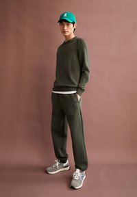 Olijfgroene sweatshirt met geribbelde ronde hals, gecombineerd met bijpassende joggingbroek. Draagt een turquoise pet met een wit icoon en grijze sneakers.