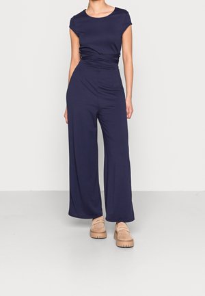 Femme portant une combinaison bleu marine à manches courtes avec jambes larges et des chaussures compensées beiges, debout sur un fond clair uni.