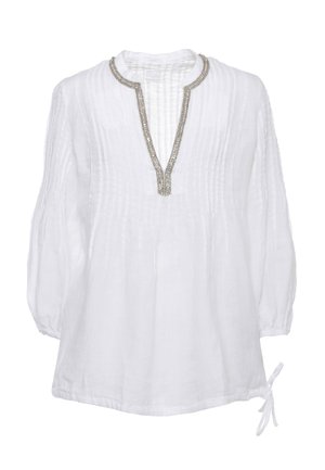Donna in piedi, indossa una camicia bianca a maniche lunghe con pieghe e scollo impreziosito da perline, pantaloni ampi celesti e sandali con punta aperta.
