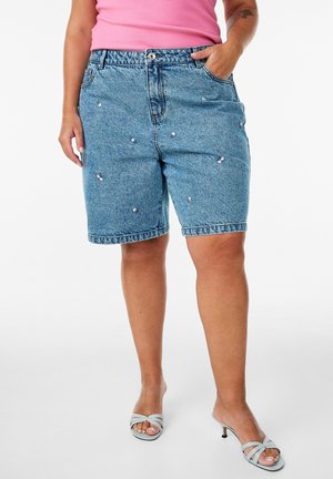 Frau trägt blaue Denim-Shorts mit kleinen weißen Verzierungen, ein pinkes ärmelloses Top und hellblaue Sandalen mit Absatz.