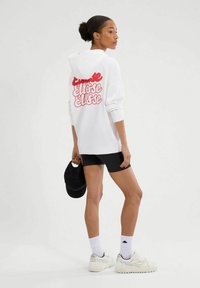 Kobieta w białej bluzie z kapturem z czerwonym napisem, czarnych szortach rowerowych, białych trampkach, trzymająca czarną czapkę, stojąca na jednolitym tle.