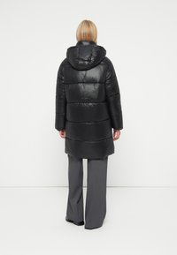 Casaco puffer preto, textura brilhante, com capuz e costuras horizontais. Combinado com calças largas cinzentas. Design simples e elegante.