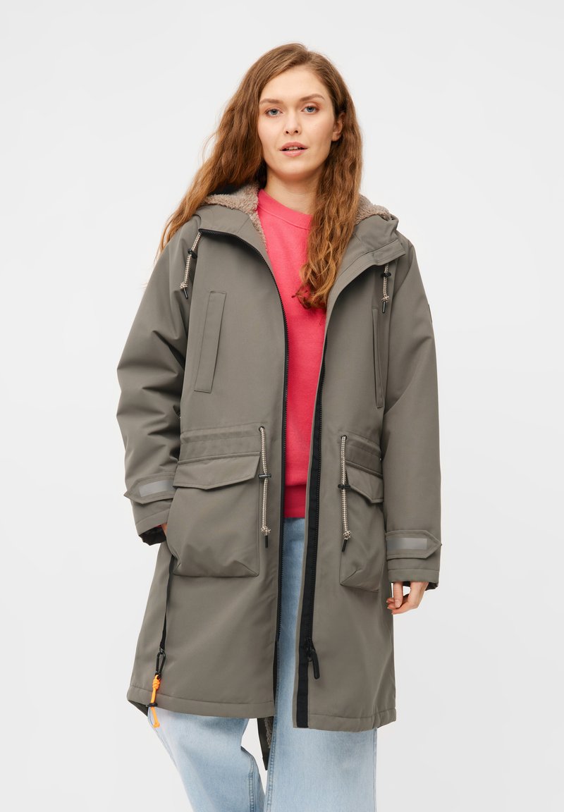 Parka grigia lunga con cappuccio foderato in pelliccia, tasche frontali e stringhe. Abbinata a una felpa rosa e jeans azzurri chiari.