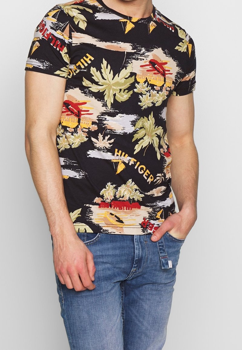 Černé tričko s krátkým rukávem s multikolorovým tropickým vzorem zahrnujícím listy, kajaky a text "HILFIGER" napříč designem.