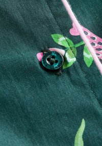 Tmavomodrý textil s ružovými a zelenými kvetinovými vzormi, obsahujúci čierny gombík so štyrmi otvormi a vyrytým nápisom „Cath Kidston, London“.