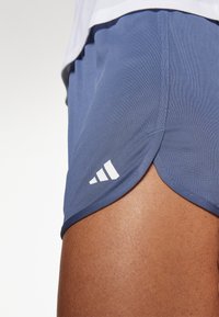 Acercamiento de una persona que lleva pantalones cortos deportivos Adidas azules con el logo en el costado y parte de una camiseta blanca visible arriba.