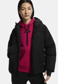 Fekete párnázott dzseki kapucnival, elülső cipzárral és oldalsó zsebekkel, világos rózsaszín kapucnis pulóver felett viselve.