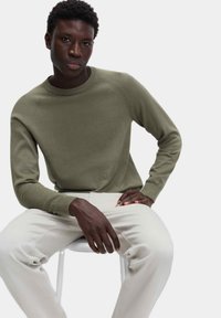 Maglione a maglia verde oliva con collo rotondo a coste e polsini, abbinato a pantaloni beige chiaro. Modella seduta su una sedia bianca.