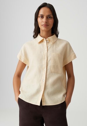 Frau mit langen dunklen Haaren trägt ein kurzes beige Hemd mit Knöpfen und dunkle Hose, steht mit Händen in den Taschen vor einem einfachen Hintergrund.