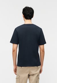 Άνδρας που φαίνεται από πίσω, φορώντας σκούρο μπλε κοντομάνικο μπλουζάκι και μπεζ παντελόνι cargo, με φόντο απλό.