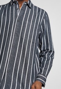 Camisa de botones en denim oscuro con rayas verticales blancas, un bolsillo en el pecho y mangas largas con puños abotonados.