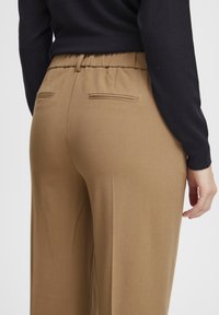 Braune, lässige Hose mit elastischem Bund, seitlichen Taschen und zwei Gesäßtaschen. Der Stoff wirkt glatt mit matter Oberfläche.