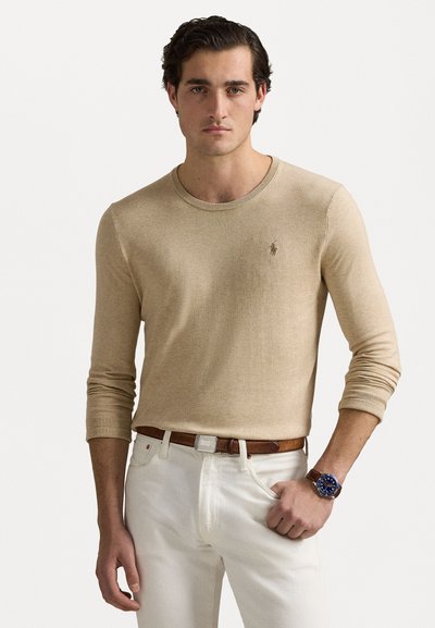 Beige dlouhý rukáv ribbed pletený svetr s kulatým výstřihem a malým logem na hrudi, doplněný bílými džíny a hnědým opaskem.