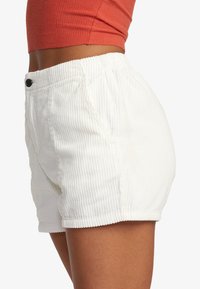 Zbliżenie na osobę w białych, żebrowanych, wysokich szortach oraz pomarańczowym, krótkim topie bez rękawów na jednolitym tle.