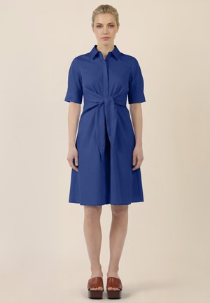 Frau, die nach vorne blickend steht, trägt ein blaues knielanges Kleid mit kurzen Ärmeln und einer vorderen Schleife, kombiniert mit braunen Plateau-Offenzeh-Schuhen.