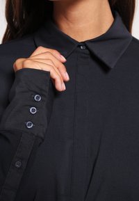 Chemise noire à col, à manches longues, avec trois boutons à chaque poignet. Tissu lisse avec une coupe structurée.
