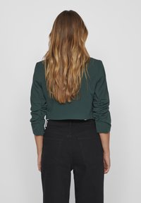 Cropped, dark green long-sleeve top with ruched sleeves, paired with high-waisted black pants, viewed from the back.