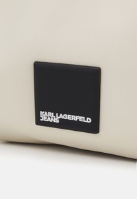 Bolso de cuero beige con una etiqueta rectangular negra con texto en relieve blanco que dice "KARL LAGERFELD JEANS." Textura suave y diseño minimalista.