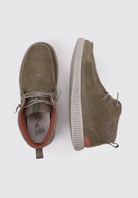 Zapatos de ante verde oliva con una suela de goma gris, que cuentan con un detalle de fieltro en el talón y ojales metálicos para los cordones. El interior tiene un forro naranja.