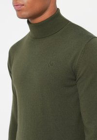 Olivno zeleni pulover s turtleneck ovratnikom, rebrasta tekstura, prilegajoča zasnova, ki ima subtilen logotip na prsih. Mehka tkanina, dolgi rokavi.
