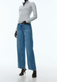 Ljust grå, texturerad stickad tröja med halvdragkedja; högmidjade, vida blå jeans; svarta sandaletter med öppen tå.