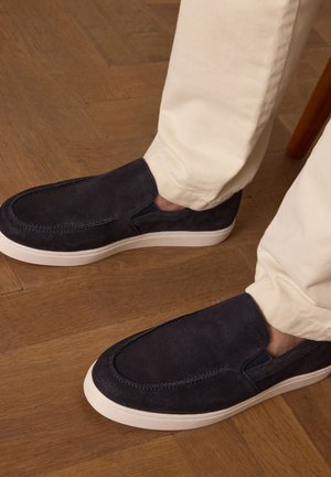 Navy blue suede slip-on shoes with a textured upper and white rubber sole, paired with cream-colored tapered pants on a wooden floor.