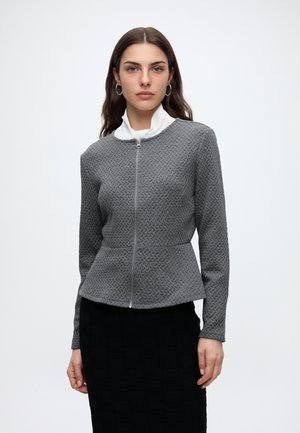 Femme aux cheveux bruns portant des boucles d'oreilles anneaux dorés, un haut blanc à col montant et une veste zippée grise texturée avec un bas à basque.