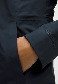 Jachetă de culoare albastră închisă cu o textură netedă; dispune de un buzunar lateral și de un logo vizibil pe manșetă într-o culoare contrastantă.