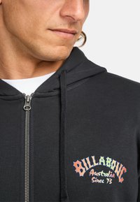 Zwarte zip-up hoodie met een kleurrijk "Billabong Australia Since 73" logo op de linkerborst en een textuurstof.