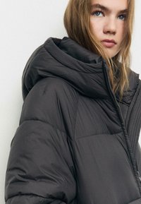 Giacca a vento nera in tonalità grigia scura con un grande cappuccio a strati. Presenta una texture trapuntata e una chiusura con zip. Il materiale morbido e imbottito mette in evidenza l'isolamento.