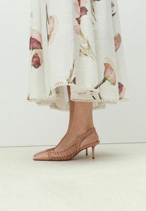 Füße in rosa gewebten Slingback-Kätzchenabsätzen kombiniert mit einem weißen Midirock mit Blumenmuster und Spitzenbesatz am Saum.