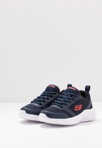 Baskets de sport navy avec tige en maille, semelle en caoutchouc blanche. Les caractéristiques incluent des lacets ronds, une languette et un logo de la marque rouge sur le côté.