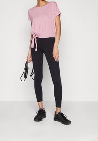 Růžové tričko s uvázáním vpředu a černé legíny, vyrobené z měkké látky, s úzkým střih a hladkou texturou. Modelka drží skákací provaz. Černé tenisky.