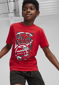 Camiseta de algodón roja con un estampado gráfico que incluye elementos en blanco y negro, como letras en negrita y un patrón circular. Se lleva con pantalones cortos negros.