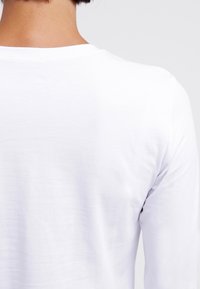 Zadní pohled na osobu s krátkými tmavými vlasy, oblečenou v prostém bílém tričku s dlouhým rukávem, na světlejším pozadí.