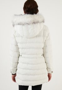 Veste blanche en duvet avec une capuche amovible bordée de fourrure, design matelassé et poches latérales. Comprend des boutons noirs aux poignets.