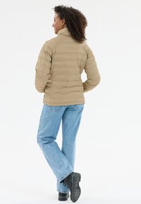 Beige dunjakke med høj krave, quiltet mønster og en slank silhuet, kombineret med lyseblå jeans og sorte snørestøvler.