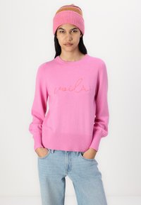 Pull en tricot rose avec "voilà" brodé en orange, doté d'un col rond et de poignets côtelés, associé à un jean bleu clair et un chapeau rose assorti.