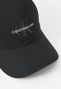 Fekete sapka sima anyagból, az elején ezüst és világosszürke színű megtűzött "CalvinKleinJeans" logóval a márkaazonosítás érdekében.