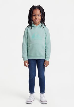 Chica con cabello largo y trenzado, vistiendo una sudadera con capucha verde menta, vaqueros azul oscuro y zapatillas blancas, posando frente a un fondo blanco liso.