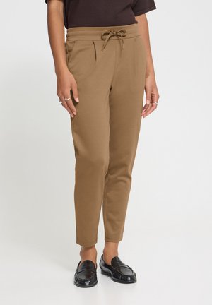 Personne portant un pantalon marron fuselé avec cordon de serrage et des mocassins noirs sans lacets sur un fond clair uni.