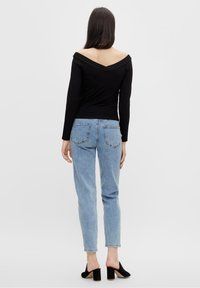 Frau mit langen, dunklen Haaren, die ein schulterfreies schwarzes Oberteil, hellblaue hochgeschnittene Jeans und schwarze High-Heel-Mules trägt und von hinten schaut.