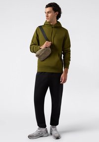 Olivovo zelená mikina s predným vreckom, čierne tepláky, béžová crossbody taška a šedé tenisky. Tkanina vyzerá mäkká a štruktúrovaná.