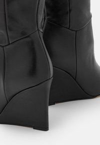 Botas negras de tacón cuña con diseño de cuero panelado, mostradas desde atrás sobre una superficie blanca con detalles de costura visibles.