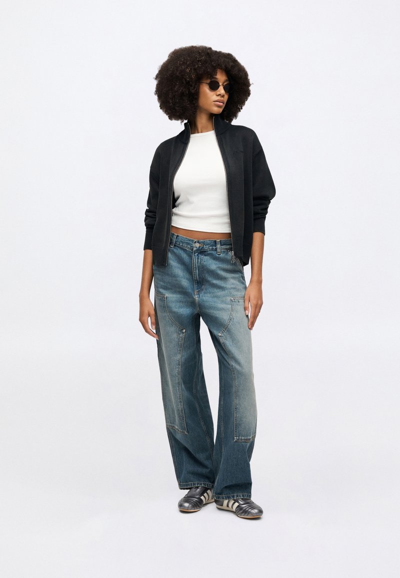 Sort bomberjakke med lynlås over en hvid croppet top, kombineret med vidde-ben, falmede blå denimjeans. Tilbehør inkluderer runde solbriller.