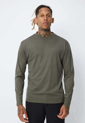 Homme avec des tatouages au cou et aux mains, portant une chemise à manches longues olive et un pantalon noir, se tenant contre un fond clair uni.