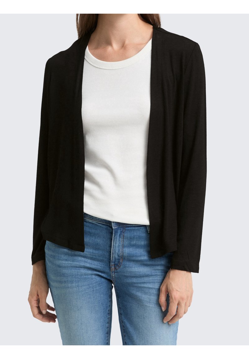 Femme portant un cardigan noir ouvert sur un top blanc côtelé à col rond et un jean denim bleu, debout devant un fond clair.
